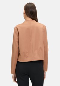 Gekürzte Jacke aus weichem, hellbraunem Stoff mit geradem Schnitt, abgerundetem Kragen und Rückenschlitz-Details, kombiniert mit schwarzen Hosen.