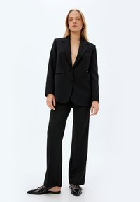 Zwarte op maat gemaakte blazer met een diepe V-hals, enkele knoopsluiting en voorzakken, gecombineerd met zwarte wijde pantalon en platte instappers.