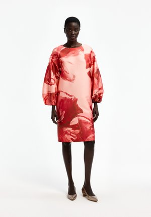 Woman stands wearing a knee-length pink dress with large red floral patterns and puffed long sleeves, paired with beige low heels.