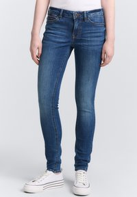 Jeans skinny de color azul con un lavado desgastado, que cuentan con cinco bolsillos, pasadores para cinturón y un cierre de botón, combinados con zapatillas blancas.