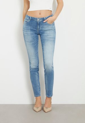 Kvinde iført lyseblå slim-fit jeans og beige spidse hæle, med den ene hånd i lommen og hvid cropped top.