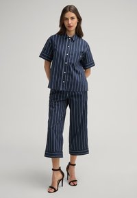 Marineblaues Set aus kurzärmeligem Hemd und cropped Hose mit weißen vertikalen Streifen. Baumwollstoff, Knopfverschluss und eine aufgesetzte Tasche am Hemd.