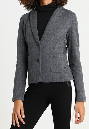 Kvinde iført en tætsiddende grå to-knaps blazer over en sort rullekrave og sorte bukser med en hvid sidestrope, står foran en hvid baggrund.