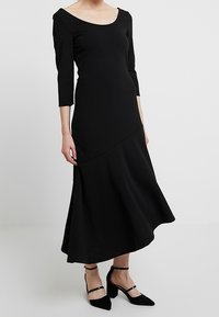 Svart, långärmad, åtsittande klänning med rundad halsringning och utsvängd nederdel. Bärs med svarta högklackade skor med ankelremmar.