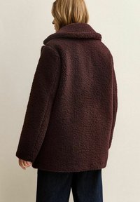 Persona vista di spalle che indossa una giacca in pile texturizzata marrone scuro sopra pantaloni scuri, su uno sfondo chiaro e uniforme.