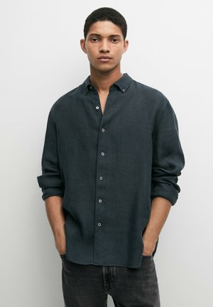 Junger Mann mit kurzen dunklen Haaren, der ein dunkelgrünes Hemd mit Knöpfen und hochgekrempelten Ärmeln sowie schwarze Jeans trägt, Hände in den Taschen.