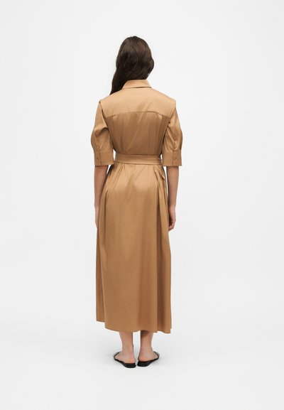 Femme aux longs cheveux foncés vêtue d'une robe midi couleur sable ceinturée, manches aux coudes et sandales plates noires, debout devant un fond blanc.