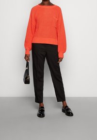 Jersey de punto naranja brillante con textura acanalada y bolsillo, combinado con pantalones tapered negros y mocasines negros. Bolso negro simple.