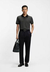 Tummanharmaa poolopaita mustalla kauluksella, yhdistetty mustiin housuihin. Vasemmassa kädessä pidetään mustaa nahkalaukkua. Kengät ovat mustat.