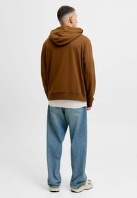 Hnědá mikina s kapucí a volným střihem v kombinaci s volnými modrými džínami a světlem barvenými teniskami. Jednoduchý design a pohodlný materiál.