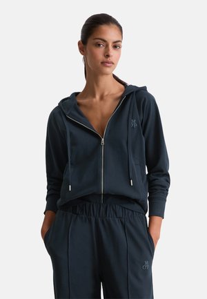 Vrouw in een marineblauwe zip-up hoodie en bijpassende broek met handen in de zakken, staand tegen een effen lichte achtergrond.