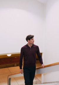 Tummanpunainen pooloneule, jossa on kolmen napin kaulus, yhdistettynä tummiin housuihin, kävelee vaaleansävyistä portaikkoa pitkin, jossa on puinen kaide.