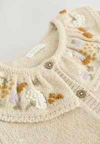 Gilet crème en tricot avec broderie florale en jaune, violet et vert. Fini avec deux boutons marron au col.