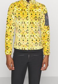 Chaqueta de forro polar en amarillo brillante con un patrón geométrico en negro y rosa, con cierre frontal de cremallera y un bolsillo con cremallera en la manga. Acabado texturizado.