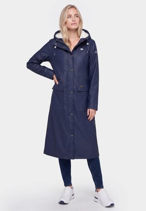 Kvinde iført en lang navy hættetrøje regnjakke med frontlommer, mørke jeans og hvide sneakers, der står med den ene hånd på hoften.