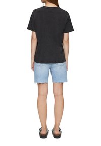 Žena stojící zády, oblečená v vypraném černém tričku, světle modrých roztrhaných džínových šortkách a černých platformových sandálech.