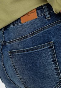 Jeans de mezclilla azul oscuro con una etiqueta de color beige claro que dice "ONLY CARMKOMA" y "Denim". Presenta un bolsillo trasero y detalles de costura visibles.