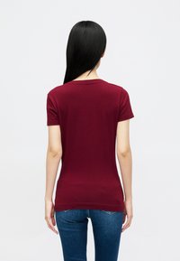Κολλητό μπλουζάκι κοντό μανίκι μπορντό, με στρογγυλή λαιμόκοψη. Λείος βαμβακερός ύφασμα, διακριτές ραφές, σε συνδυασμό με μπλε τζιν παντελόνι.