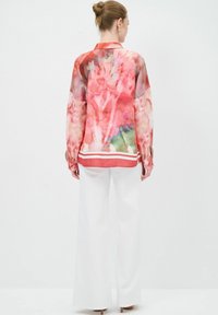 Camicia a maniche lunghe, trasparente, con un motivo floreale nei toni del rosa e del verde. Abbinata a pantaloni bianchi a gamba larga, con un orlo a righe.