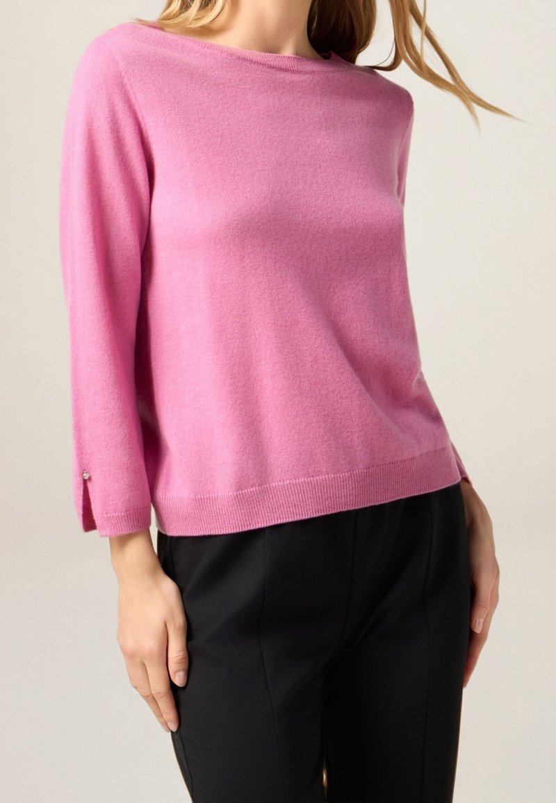Femme portant un pull rose à manches longues avec détail de boutons au poignet et un pantalon noir sur un fond neutre.