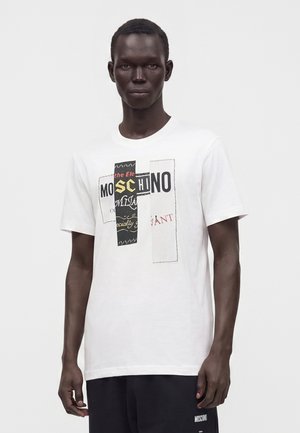 Mand iført hvid Moschino t-shirt med sort, gul og rød grafisk tekst stående mod en ensfarvet hvid baggrund.