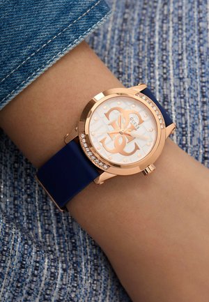 Montre en or rose avec un cadran rond, un cadran blanc à motifs, un bracelet en silicone bleu marine, et rehaussée de cristaux autour de la lunette.