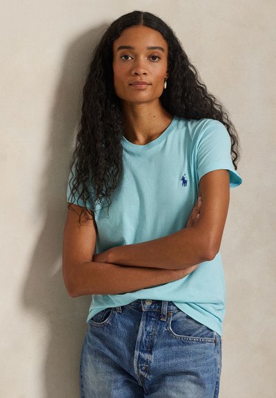 Ανοιχτό μπλε βαμβακερό μπλουζάκι με στρογγυλή λαιμόκοψη και κοντά μανίκια, με μικρό μπλε λογότυπο στο στήθος, συνδυασμένο με μπλε τζιν παντελόνι.