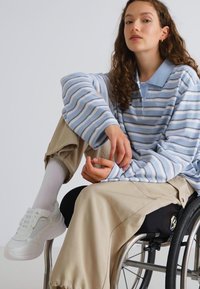 Chemise polo rayée bleu clair, pantalon large beige, baskets blanches et un fauteuil roulant avec un coussin noir, présentant un logo sur le côté.