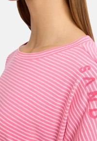 Rosa randig skjorta med rund halsringning och mjuk bomullstextur. Ordet "SAND" är tryckt med feta röda bokstäver på ärmen.