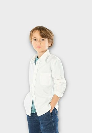 Chemise blanche à boutons avec les manches retroussées et une poche poitrine, superposée sur une chemise bleu-vert. Associée à un jean bleu foncé.