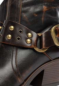 Stivaletto in pelle nera con cinturino in pelle marrone, rivetti in ottone e accessori metallici. Presenta una superficie testurizzata e un tacco in legno.