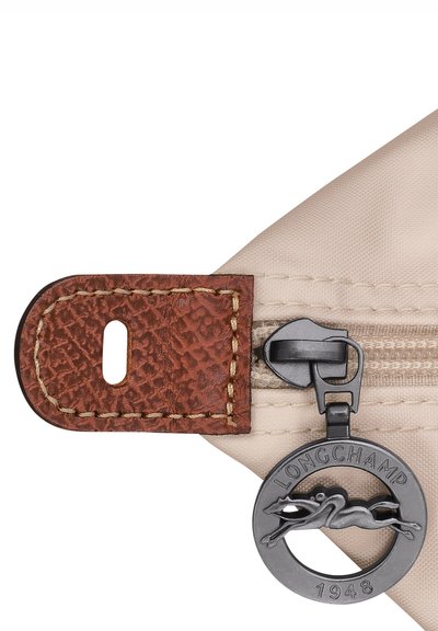 Beige nylon bag corner featuring a textured brown leather tab, gold stitching, black zipper pull, and a circular metal logo charm.
