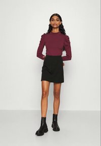 Top de manga longa em bordeaux com ombros volumosos, combinado com uma saia mini preta e botas pretas de tornozelo, contra um fundo neutro.