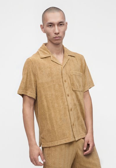 Homme portant une chemise marron clair à manches courtes boutonnée et un pantalon assorti avec un motif à rayures verticales texturé.