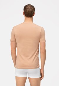 T-shirt aderente beige chiaro realizzata in cotone morbido, con maniche corte e scollatura rotonda. Indossato con boxer bianchi.