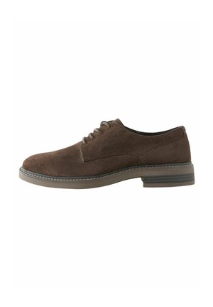 Zapato de vestir de ante marrón con puntera redondeada, diseño con cordones y una suela de color claro en contraste con un patrón de tracción sutil.