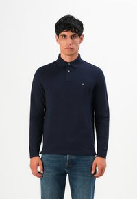 Vyras stovi sukęs į priekį, vilkintis tamsiai mėlyną ilgomis rankovėmis polo marškinėlį ir mėlynus džinsus prieš paprastą baltą foną.