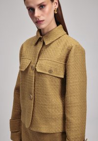 Giacca strutturata in giallo senape con texture, colletto, tasche anteriori e bottoni rotondi dorati. Presenta una vestibilità aderente e maniche lunghe.