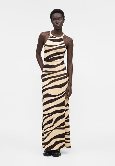Γυναίκα φοράει αμάνικο, μακρύ φόρεμα με μπεζ και μαύρες ζεβρέ ρίγες, σε συνδυασμό με μαύρα παπούτσια, στέκεται μπροστά σε λευκό απλό φόντο.