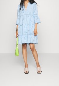 Ljusblå bomullsklänning med broderade detaljer, V-ringning, utsvängda ärmar och flerlagat volangfåll. Kombinerad med grön väska och nude blockklack sandaler.