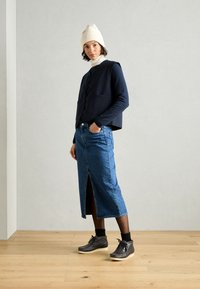 Vrouw in een crèmekleurige beanie, marineblauwe vest over een marineblauw longsleeve, blauwe denim midirok met een split aan de voorkant, zwarte sokken en grijze veterschoenen.
