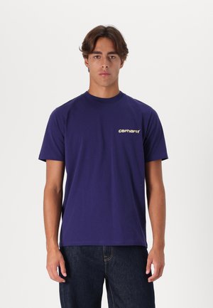 Camiseta de algodón morada con cuello redondo, mangas cortas y un pequeño estampado gráfico en el lado izquierdo del pecho. Combinada con jeans oscuros.
