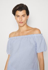 Blusa off-the-shoulder com riscas verticais azuis e brancas, tecido texturizado e mangas bufantes. O decote com babados acrescenta detalhe.