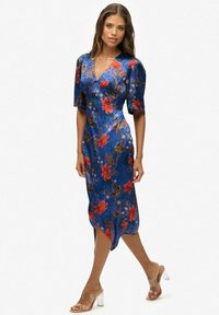 Blauwe bloemenjurk met korte pofmouwen, met rode bloemen en groene bladeren, een diep V-hals, en een zijde-split; gecombineerd met schoenen met transparante hak.