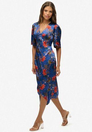 Blauwe bloemenjurk met korte pofmouwen, met rode bloemen en groene bladeren, een diep V-hals, en een zijde-split; gecombineerd met schoenen met transparante hak.