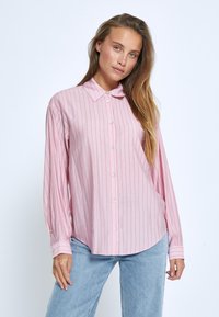 Camicia a maniche lunghe di colore rosa chiaro con righe bianche verticali, chiusura con bottoni, colletto a punta e fondo arrotondato. Abbinata a jeans azzurri.