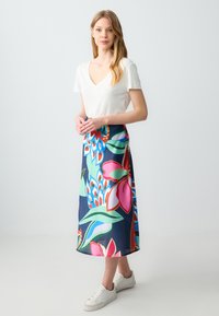 Lange rok met bloemenpatroon in marineblauw, voorzien van grote roze en groene bloemen; gecombineerd met een witte V-hals t-shirt en witte sneakers.