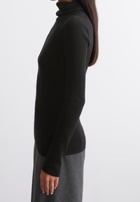 Pull à col roulé côtelé noir avec une coupe ajustée, manches longues et texture douce, porté avec une jupe grise contrastante.