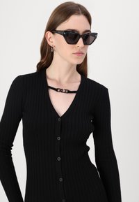 Fekete bordázott kardigán V-nyakkal és teljes hosszúságú ujjal, gombos záródással és egyedi fekete pánt részlettel a dekoltázs mentén.