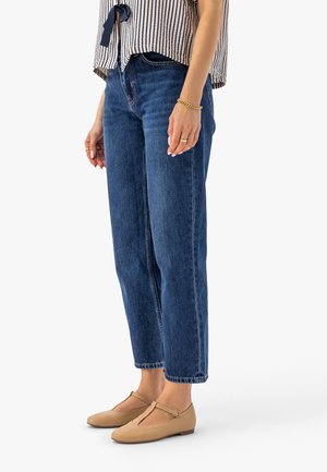 Kobieta w niebieskich dżinsach z wysokim stanem, beżowych balerinach na pasku w kształcie litery T, bluzce w paski z wiązaniem z przodu, złotej bransoletce i pierścionkach, stojąca na białym tle.